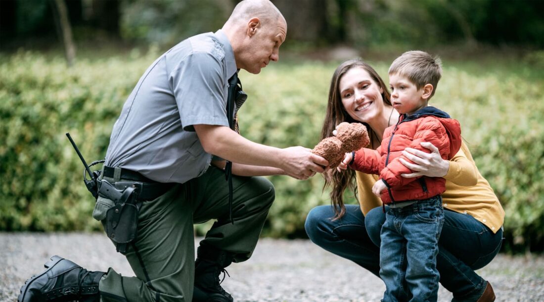 Police officer kneeling down handing a teddy bear to little boy with his mom